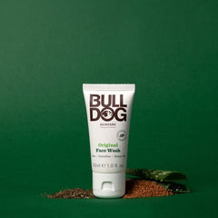 A tube of Travel Original Face Wash stands upright on a green background, with small piles of seeds and a green patterned cloth nearby, showcasing mens skincare made with natural ingredients.