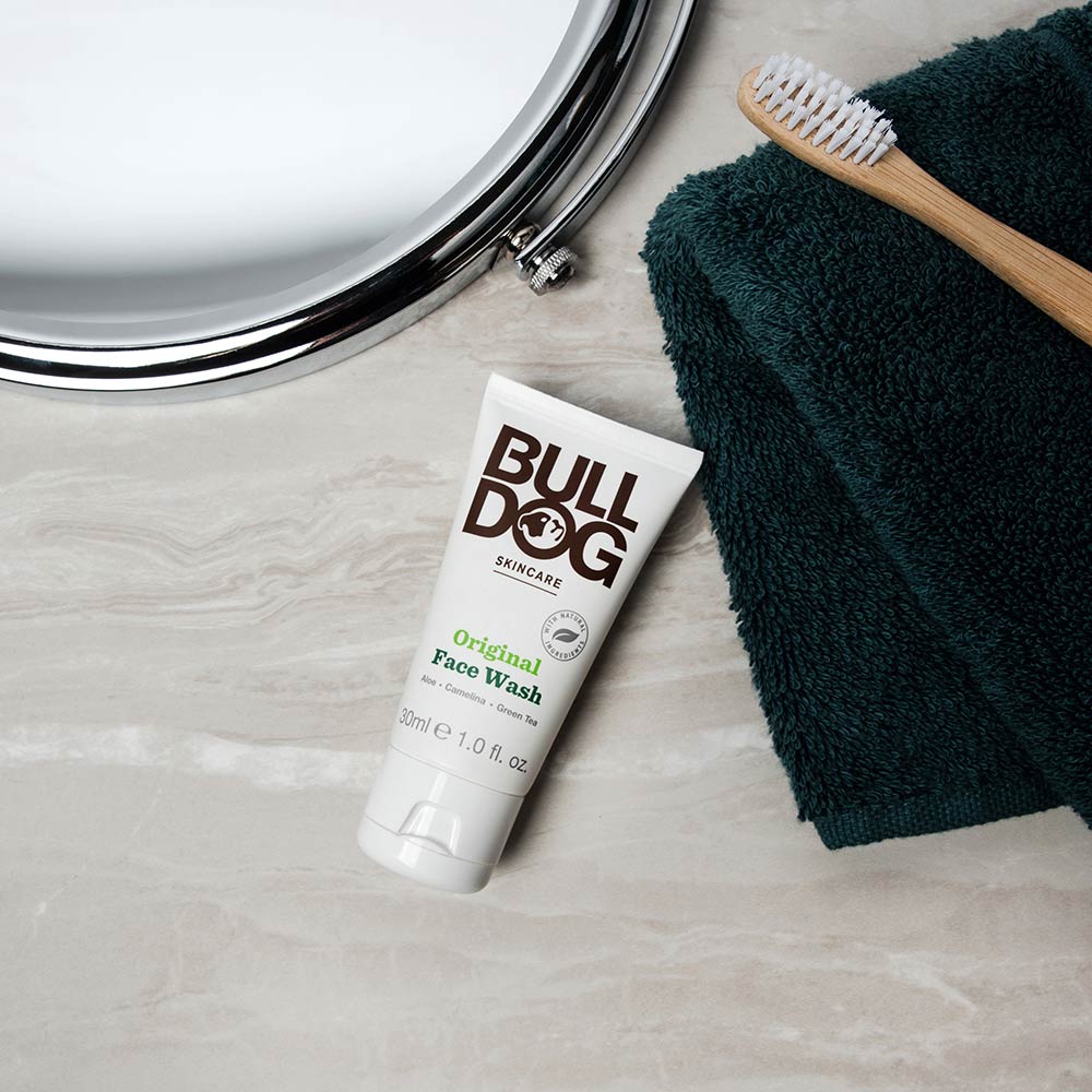 A tube of Travel Original Face Wash rests on a marble countertop next to a dark green towel, a wooden toothbrush, and the edge of a round mirror.