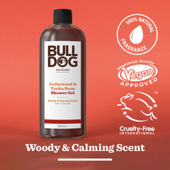 A bottle of Cedarwood & Tonka Bean Shower Gel sits on a red background, labeled 100% natural fragrance, vegan approved, and cruelty-free. Text reads: Woody & Calming Scent.