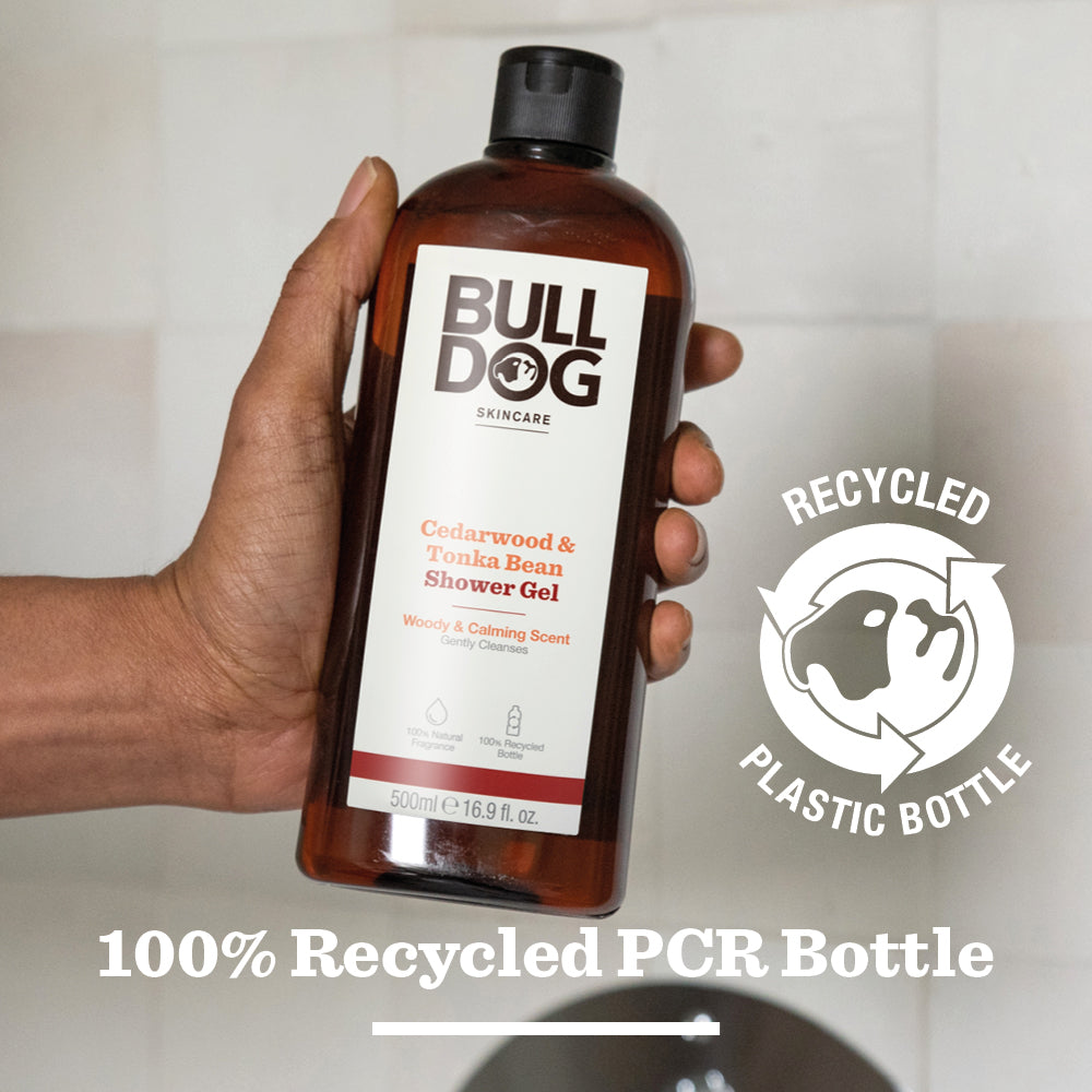 A hand holds a 500ml bottle of Cedarwood & Tonka Bean Shower Gel, featuring a woody natural fragrance. The bottle is made from 100% recycled PCR plastic, and a recycling symbol is visible on the packaging.