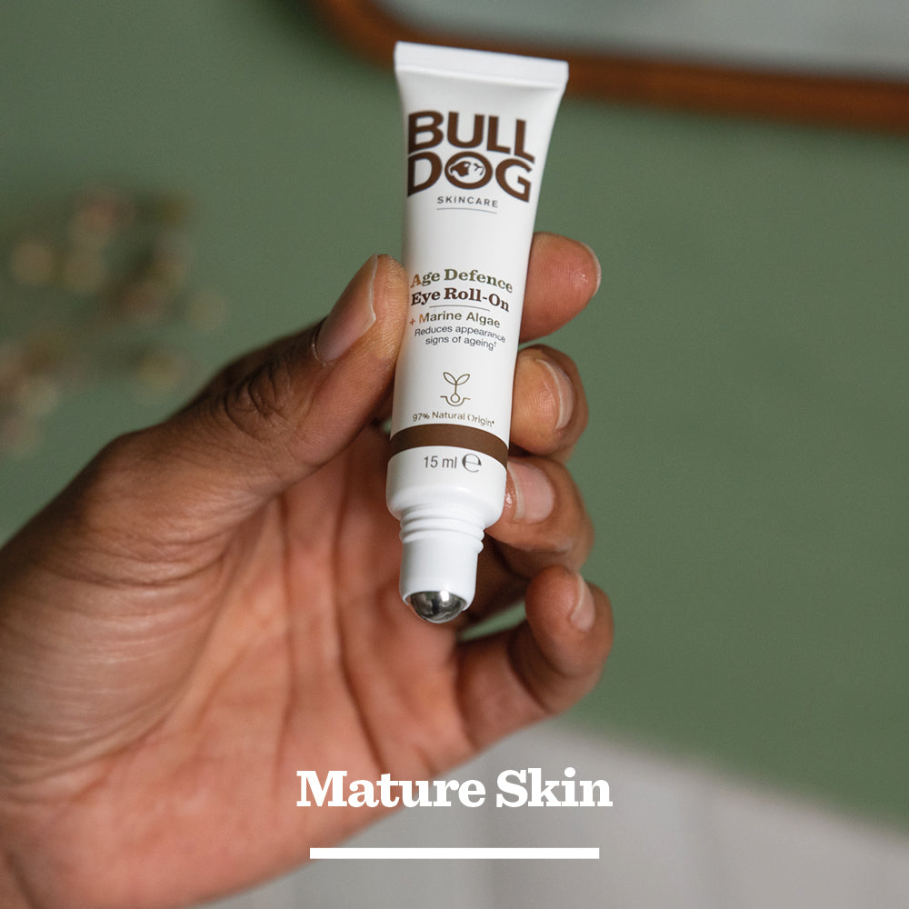 A hand holds Age Defence Eye Roll-On, a men’s skincare product with a metal roller tip and marine algae ingredient, set against a blurred green background.