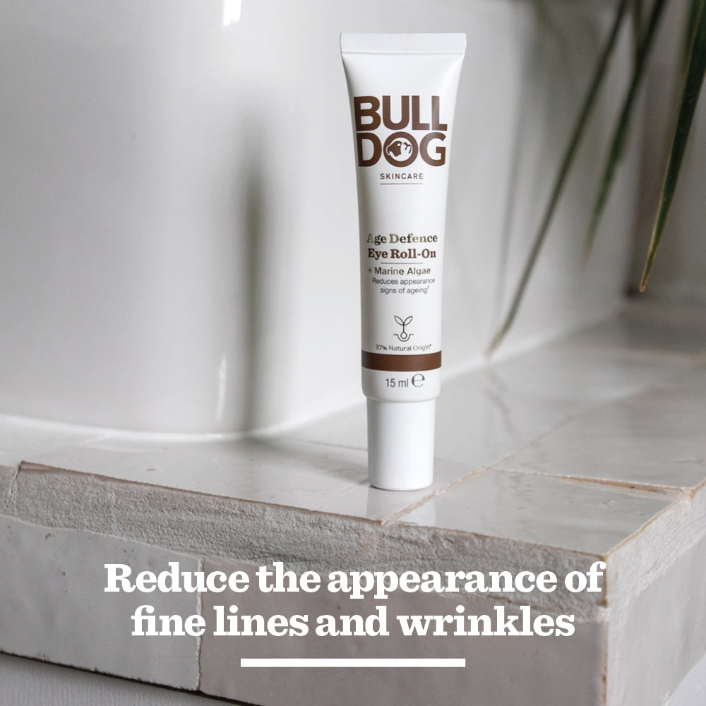A tube of Age Defence Eye Roll-On rests on a tiled surface, text below says Reduce the appearance of fine lines and wrinkles. Green plant leaves are in the background.
