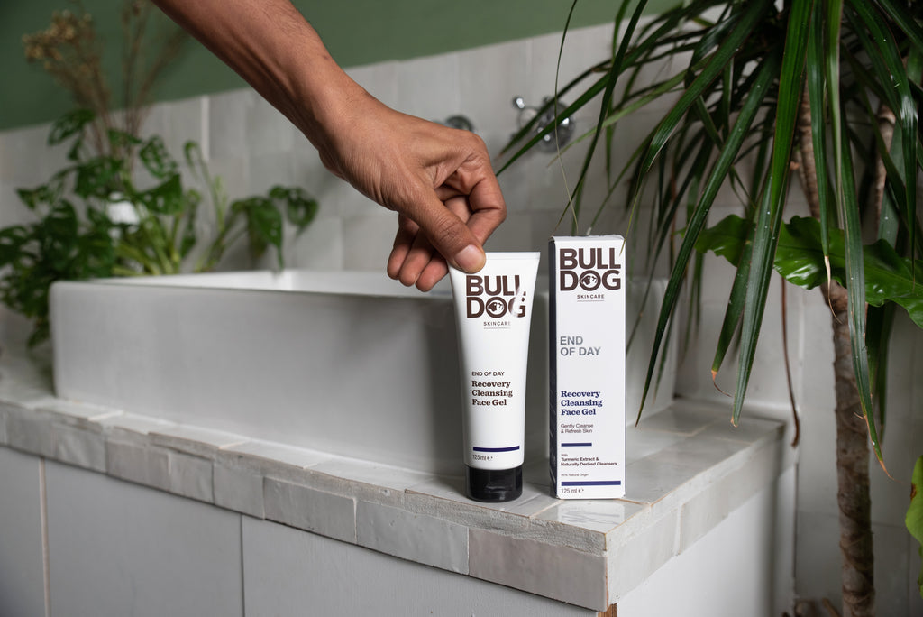 A hand picks up a tube of End of Day Recovery Cleansing Face Gel next to its box, both placed on a bathroom sink with green plants in the background.