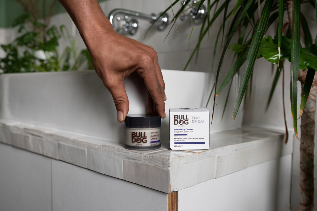 A hand reaches for a jar of End of Day Recovery Cream, placed on a bathroom counter beside its box, with green plants and a white sink in the background.