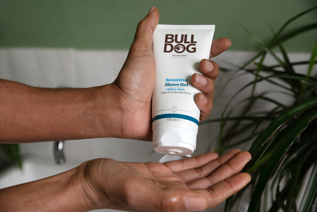 A person dispenses Sensitive Foaming Shave Gel from a white tube into their palm, with green leaves and a white sink in the background, evoking care for sensitive skin.