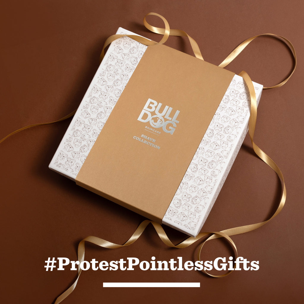 The Premium Gift: Shave Collection, featuring a bamboo razor, is wrapped in a brown paper sleeve with gold ribbon and displayed on a brown surface. White text below reads: #ProtestPointlessGifts—perfect for men who value quality.