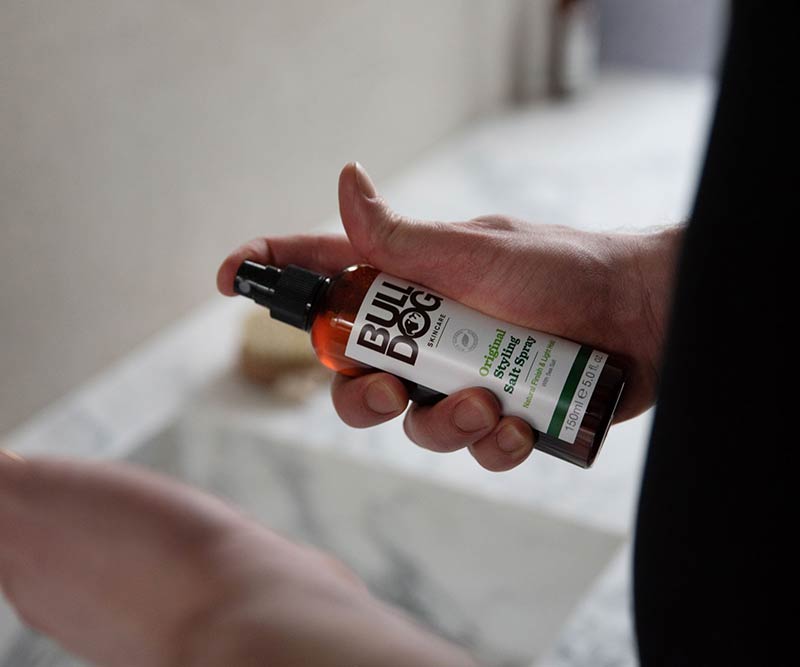 A person holds a bottle of Original Hair Styling Salt Spray—made with naturally derived ingredients—over a bathroom sink, preparing to use the product.